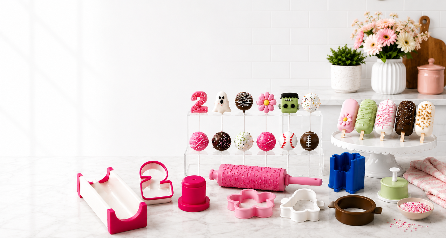 A colorful assortment of baking and dessert-making tools arranged on a white marble surface, including pink and white cookie cutters, a pink rolling pin, a pink number 2 mold, chocolate-covered popsicles, and pink sprinkles, with flowers and shelves in the background.